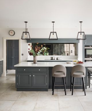 classic style kitchen with white countertops, gray cabinets, pale gray tiles, bar stools, metal and glass pendant lights, mirror, vase of flowers,