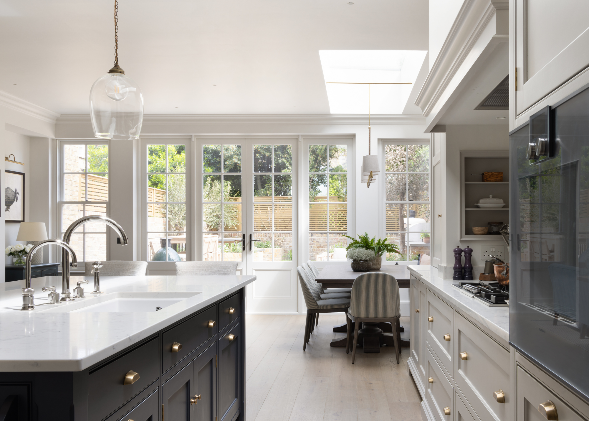 A navy blue and neutral colour spacious kitchen with white worktops and a dining table at the end looking out into the garden
