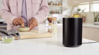 The Sonos Era 100 in black on a kitchen work surface as someone prepares food in the background