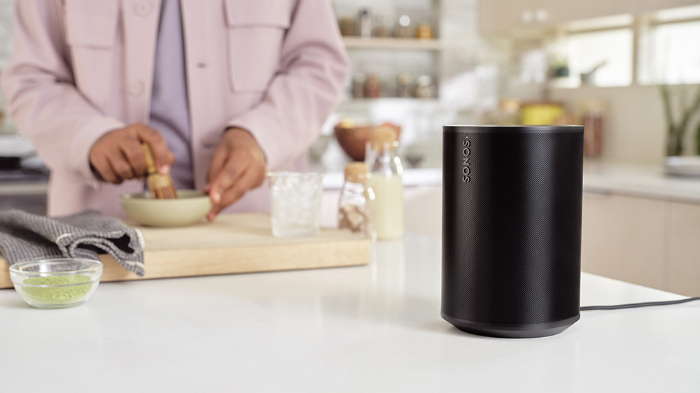 The Sonos Ear 100 in black on a kitchen work surface as someone prepares food in the background