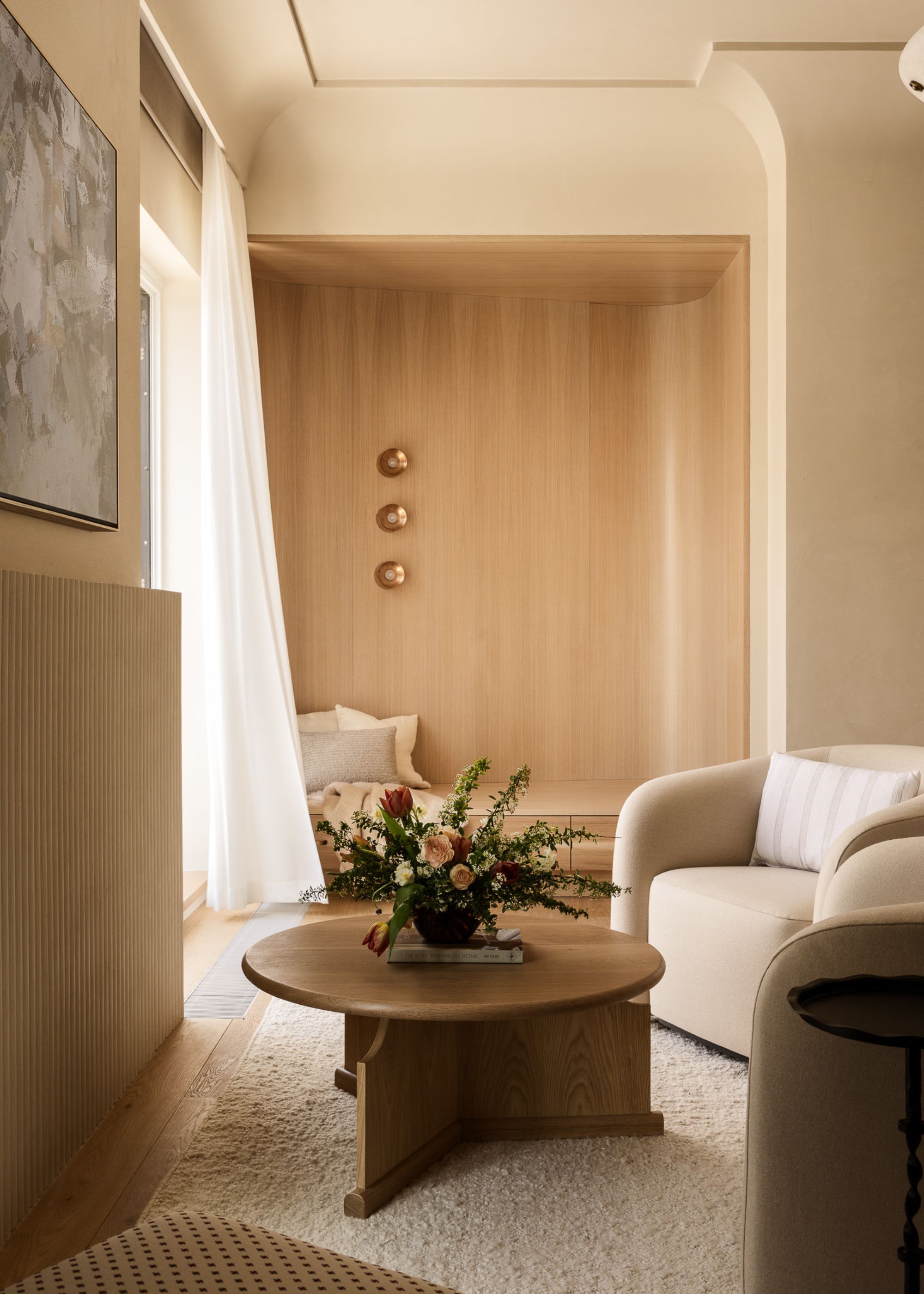 A curved, built-in niche in a modern bedroom is clad in pale wood for a cozy reading nook, with three small wall lights, behind a small seating area with two cream armchairs in front of a round table