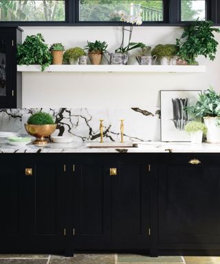 black and white kitchen with marble counterops, black units, brass handles and faucet, white floating shelf with plants, artwork