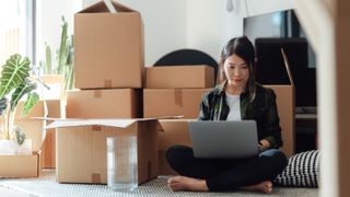 Asia woman sat on floor with laptop with piles of brown boxes behind her