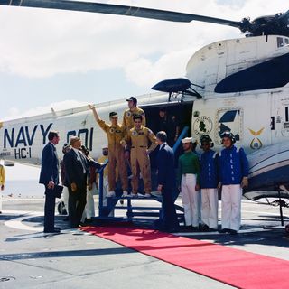 The three apollo 17 crewmembers emerge from a large helicopter and acknowledge the welcome committee. A red carpet leads to the helicopter doors.