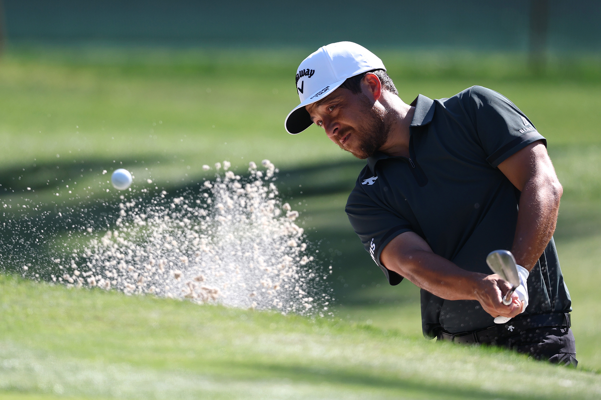 Xander Schauffele hitting a bunker shot with sand splashing up around him