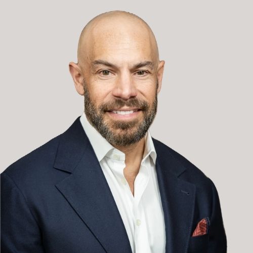Image of a man in a white button-down and a navy blue blazer in front of a beige background.