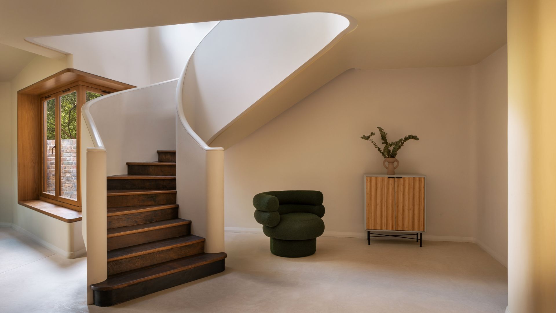 A minimalist home with a smooth curved staircase by a stacked green armchair and a wood sideboard with a vase of stems