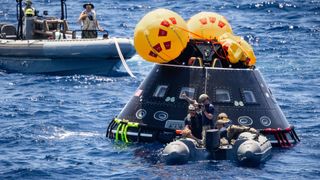 a black cone-shaped spacecraft floats in the water as people on rafts around it tie ropes to it