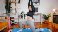 woman performing a runner's lunge with arms extended upward in a living room setting on a blue exercise mat