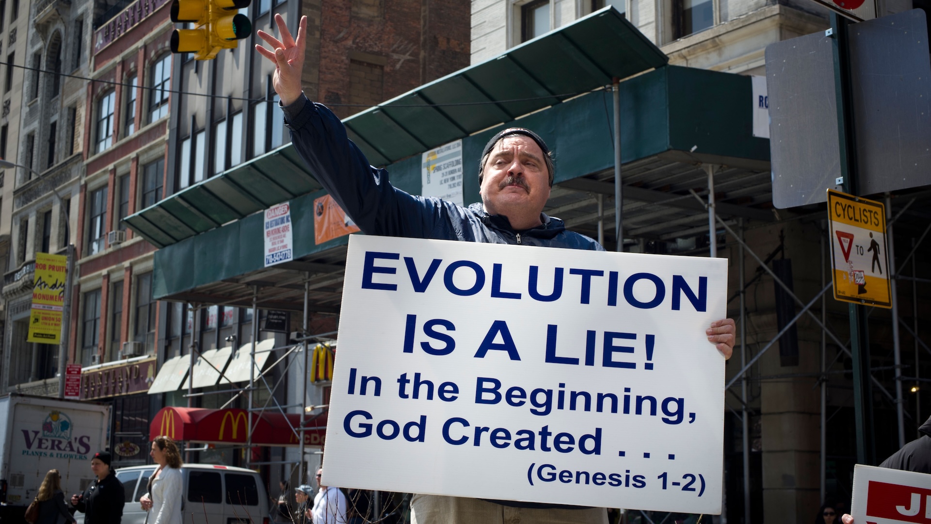 a protestor holds up a sign that reads "Evolution is a lie! In the beginning, God Created... (Genesis 1-2)"