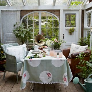 conservatory with floorboards, white doors, dining table with a green and white tablecloth with a rose pattern, a blue wicker chair and brown cushioned chair