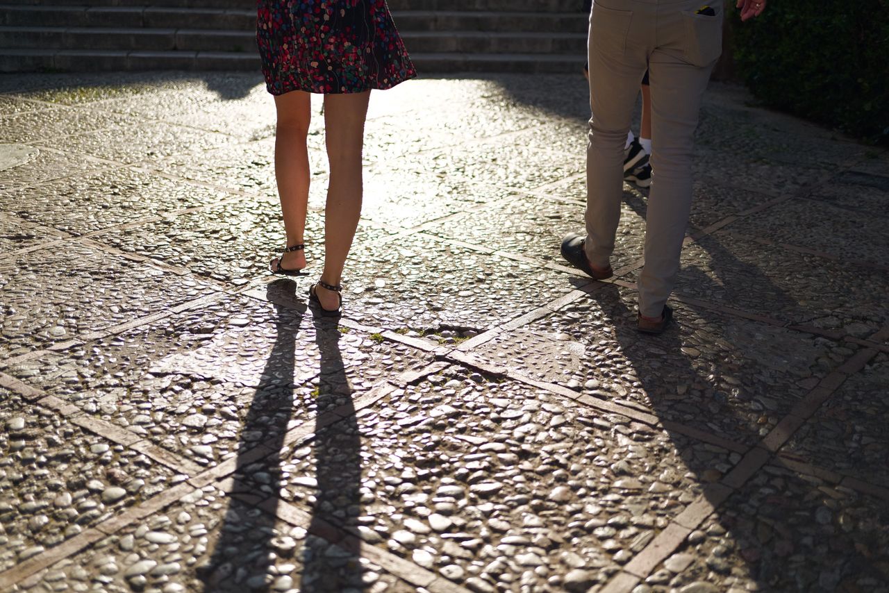 Two people walking across sunlit cobblestones, their long shadows stretching across the textured pavement.