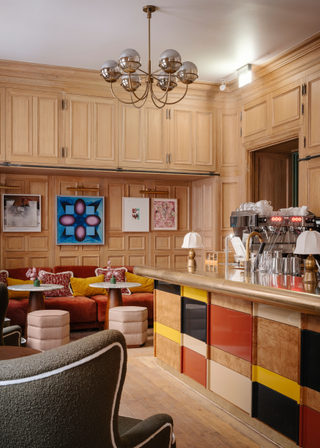 A library bar in a members' club features a blocky wooden bar counter with brass details, art deco references, and colorful framed artworks.