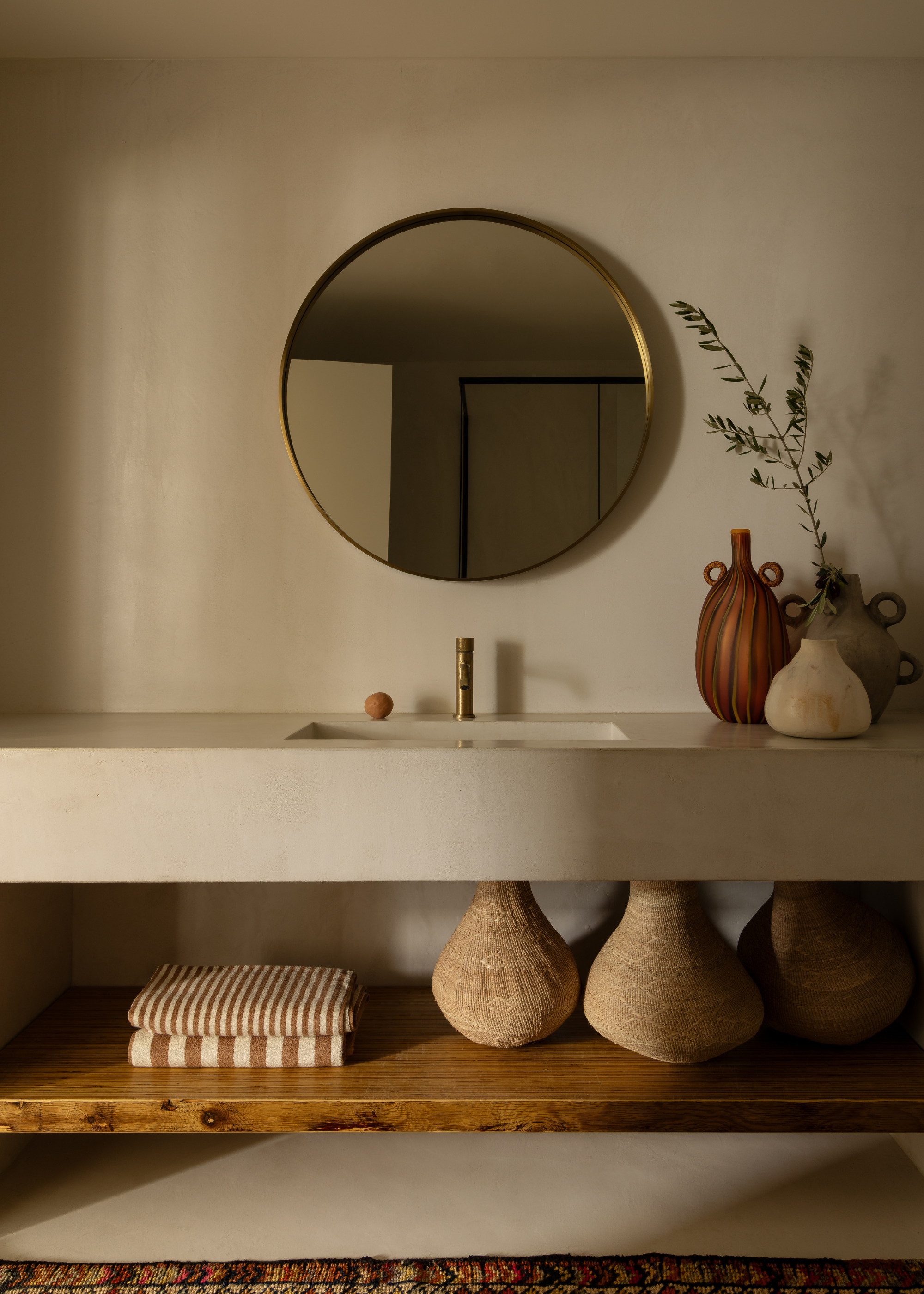 A minimalist earthy bathroom with a round mirror, a trio of vases, a wooden shelf with striped towels and woven baskets