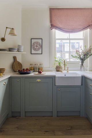 Image of a white kitchen with blue cabinets and a white countertop. There is a window above the sink that has a chic, red curtain blind halfway drawn.