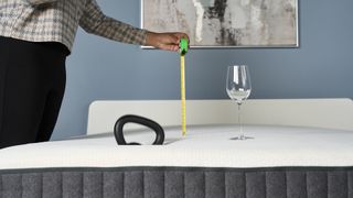 A wine glass stands upright on the Emma Elite Mattress, with a weight dropped on the surface from a short distance away. A hand holds a measuring tape showing the distance of the weight drop