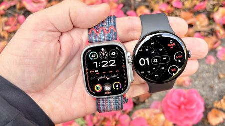 Close-up of the Apple Watch Ultra 3 and Google Pixel Watch 4 in a user's hand against a colorful out-of-focus background