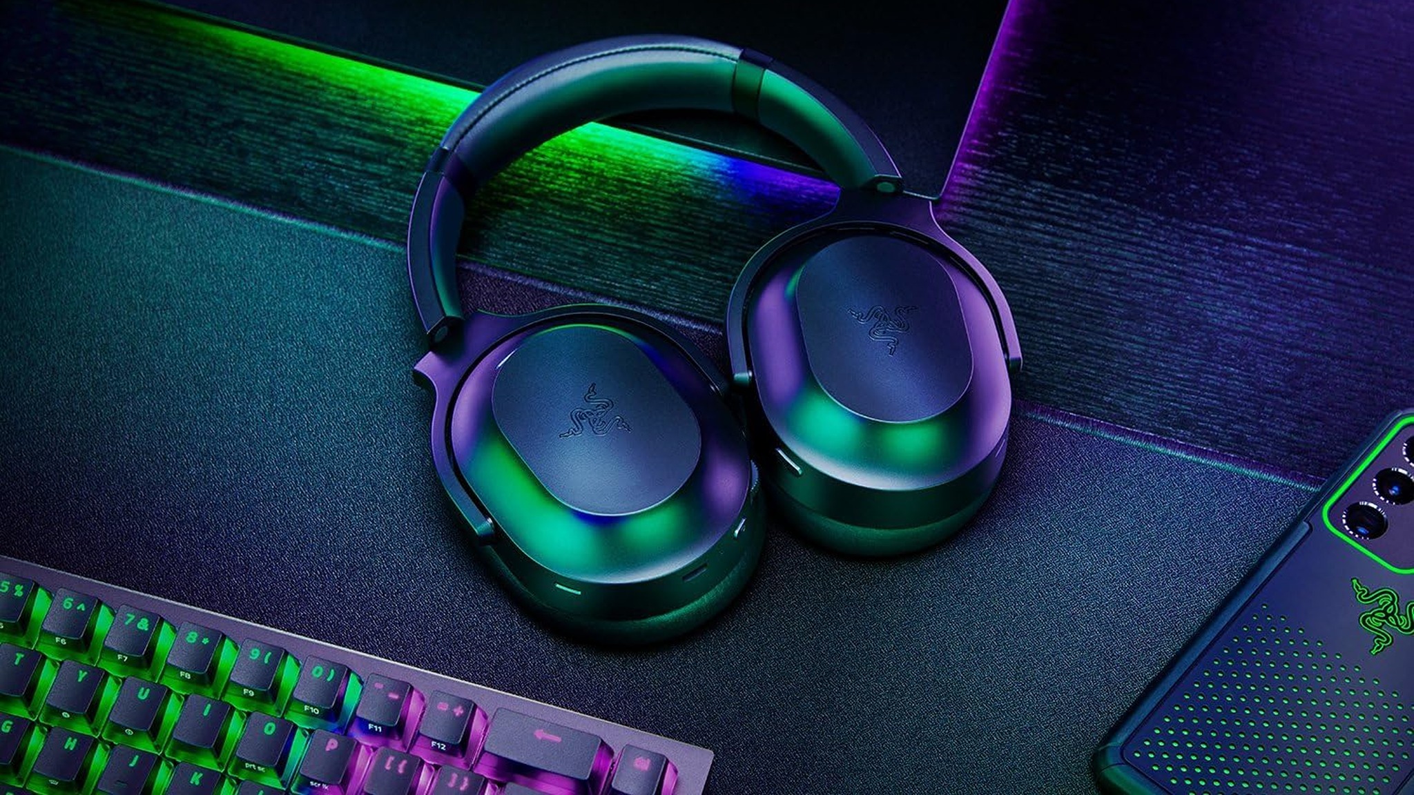 Sleek gaming headphones in green and purple hues sit on a dark desk, surrounded by a glowing RGB keyboard and a smartphone, creating a vibrant tech ambiance.