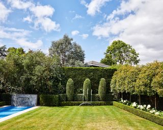 A lawn next to a swimming pool and surrounded by hedges, trees and flowerbeds