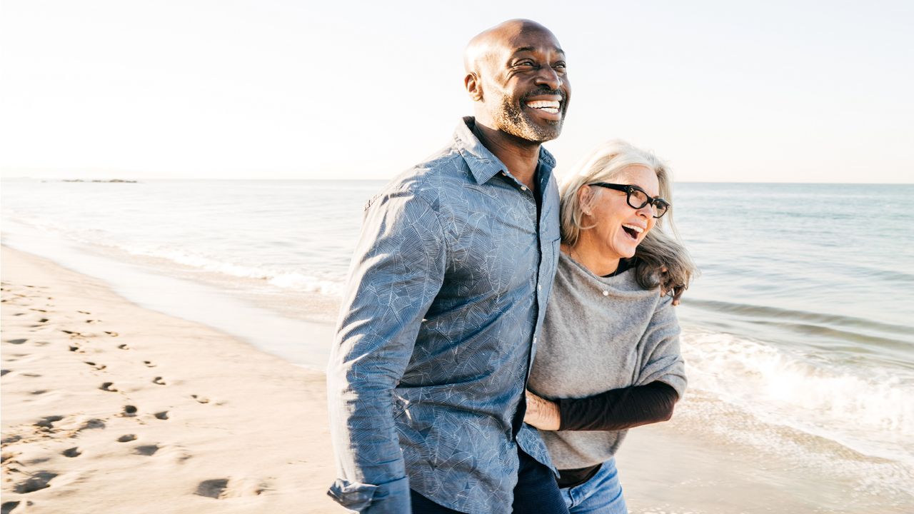 An older couple smile and laugh as they walk arm in arm on the beach.