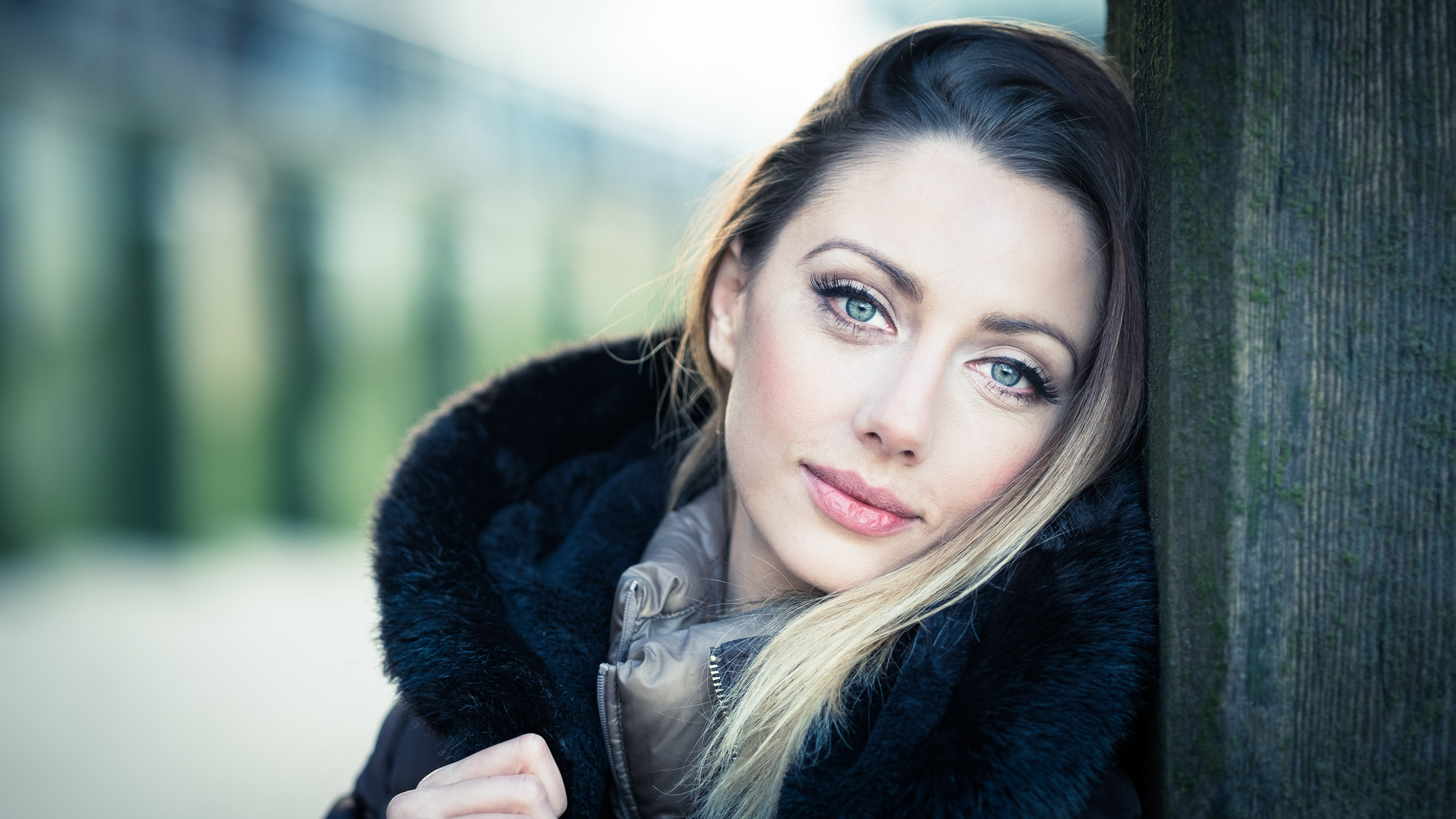  A young woman with long blond hair and blue eyes is leaning against a wooden post outdoors.