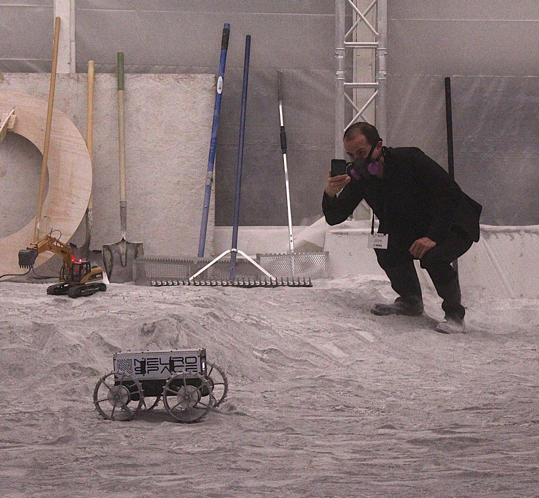 A man wearing all black with a black mask takes a photo of a small lunar rover