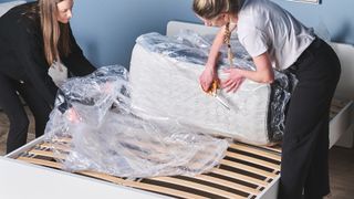 Two mattress testers using scissors to take off plastic around a mattress on a bed frame