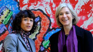 A woman with short curly black hair stands next to a slightly taller woman with straight grey hair in front of a colorful mural