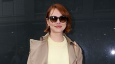 Emma Stone wearing a butter yellow sweater, a white slip skirt and a beige trench coat, carrying a Louis Vuitton bag