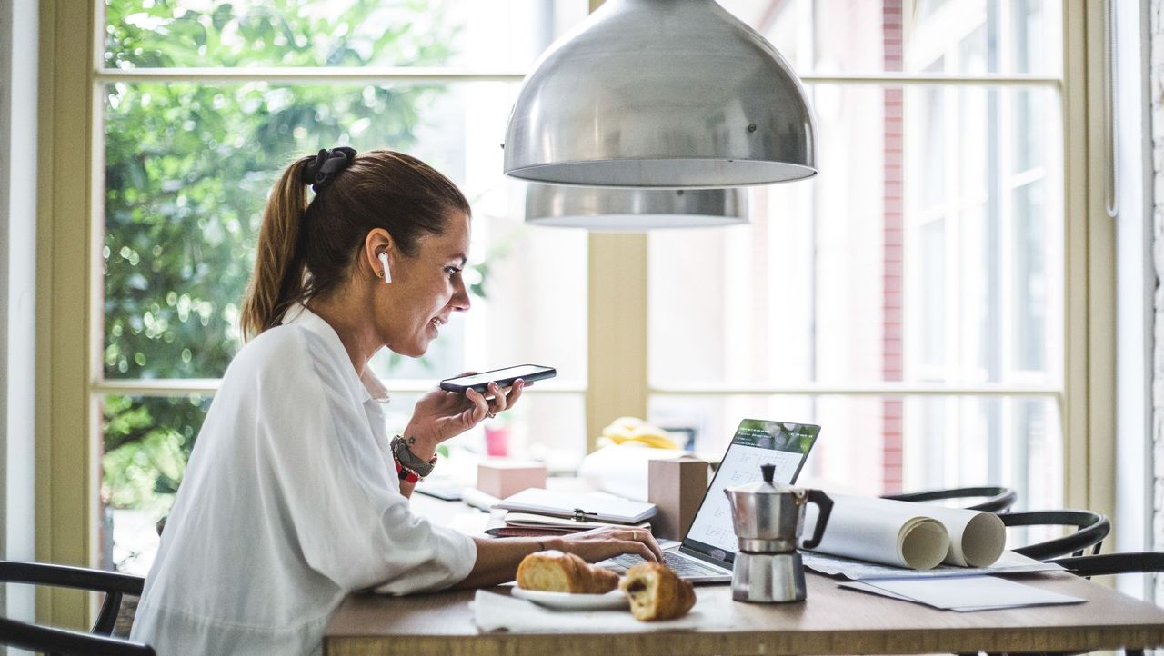 smiling female entrepreneur using laptop while talking through smart phone at home office