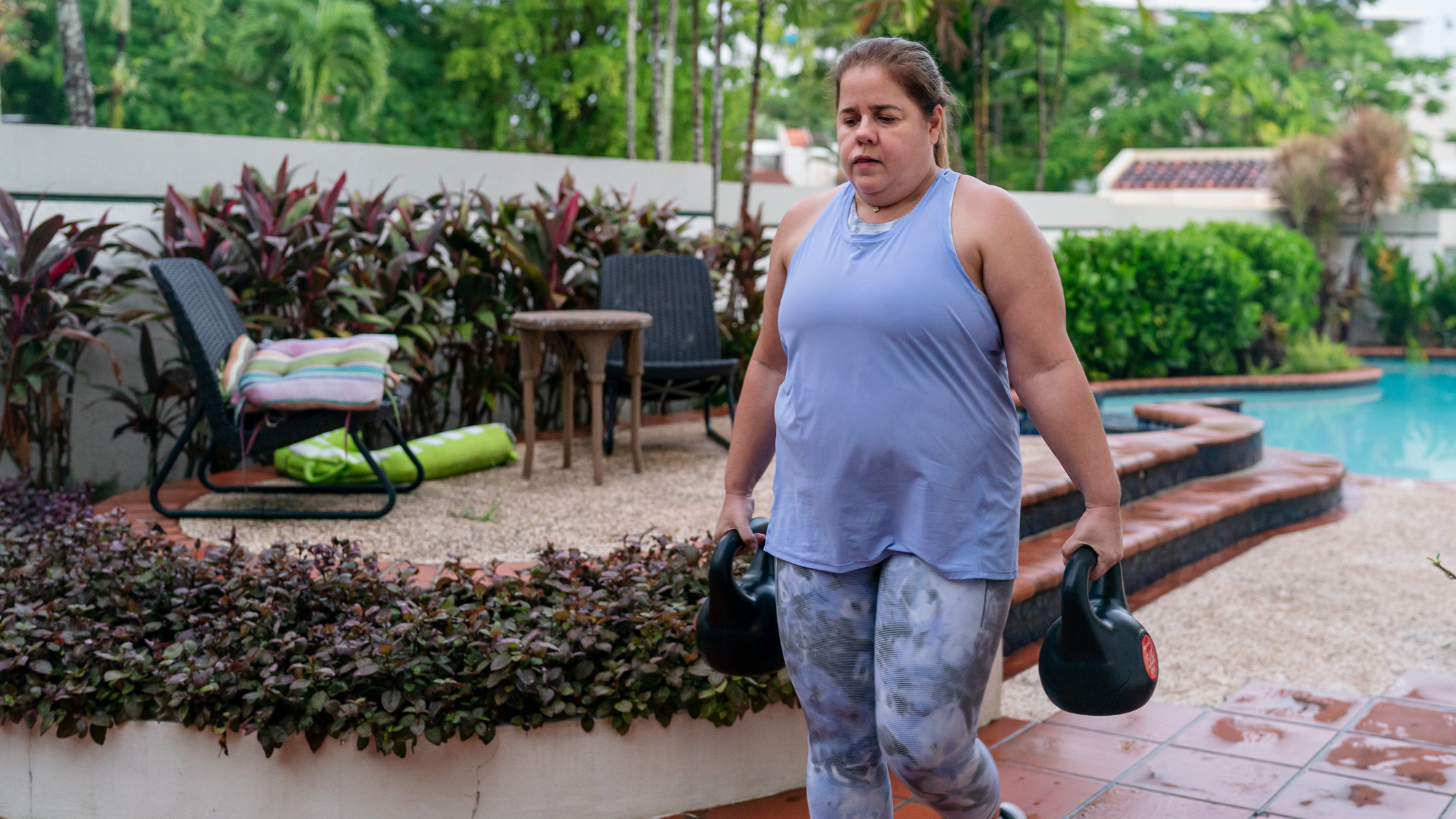 Woman carries kettlebells by her side in a domestic garden with a pool in the background
