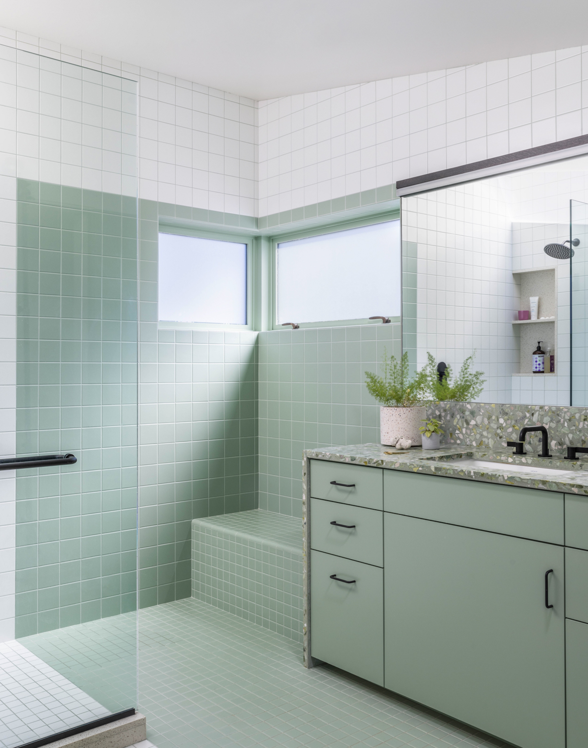 A mint green bathroom with square wall tiles that blend seamlessly into the floor tiles. The sink areas work top is a speckled mint green stone with black tapware and mirror above.