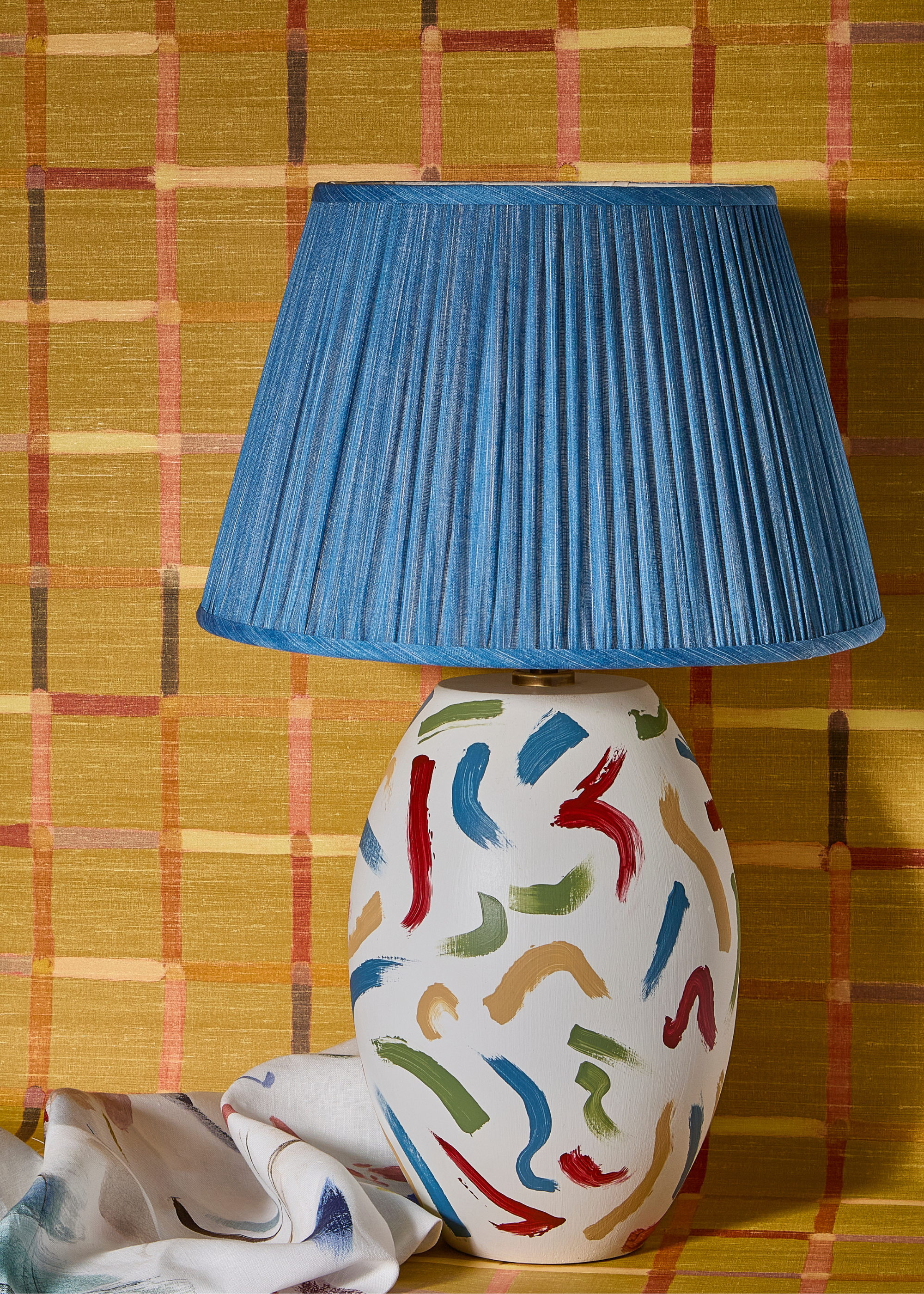 a table lamp with a blue lamp shade and a white base with different coloured squiggly lines that have been visibly painted on next to a similar designed scarf scrunched up next to it against a vibrant yellow background with cross-crossing lines