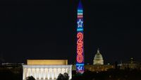 A vertical building is lit up in red, white and blue colors with the numbers 250 on it from top to bottom next to a white illuminated building with columns against a dark night sky.