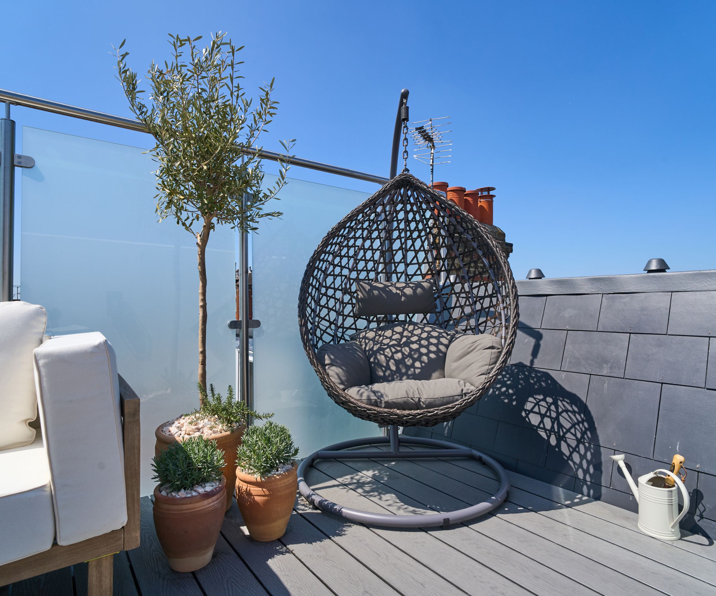 Rooftop terrace with hanging egg chair, potted plants, slate cladding and frosted glass balustrade under a clear blue sky.