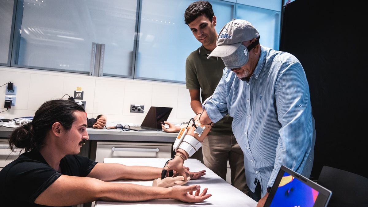 An older man in a ballcap, button-down shirt and blindfold wears a prosthetic hand on his right arm. Two scientists dressed in casual clothing watches as the man touches one of the scientist&#039;s arms