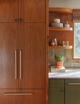 Image of a kitchen that has a warm wood built-in pantry. The cabinets are sage green with terrazzo countertops. The cabinets have golden pulls and there are a few decorations on the countertops including a stack of books and a potted plant.