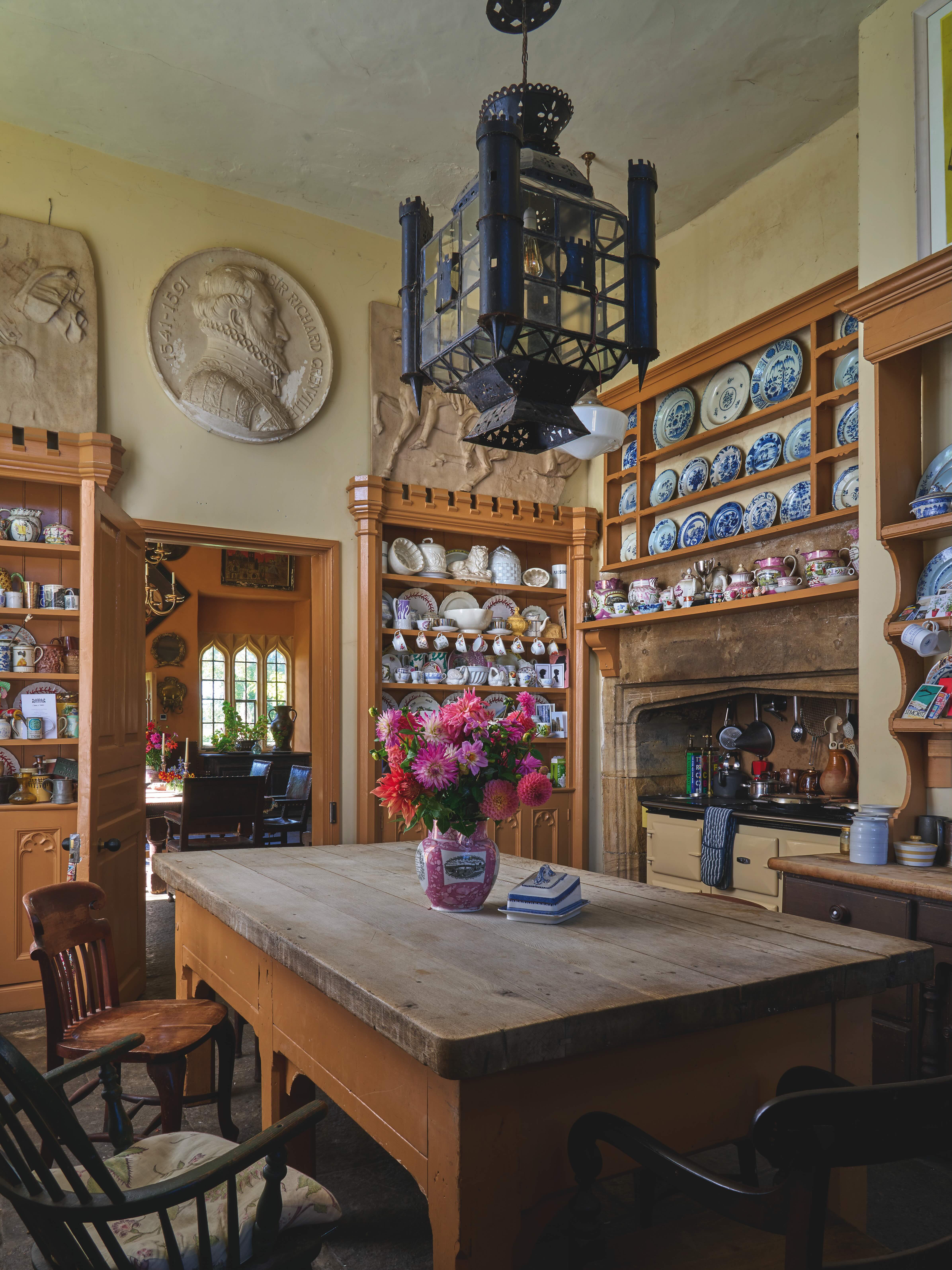 The kitchen. Above a large wooden table is a cast-iron lamp, reliefs can be seen on the walls and dressers are packed with plates.
