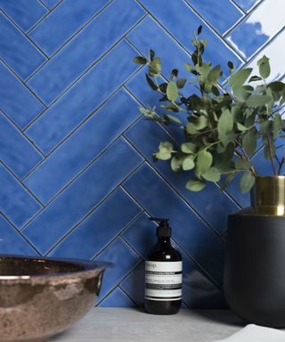 A wall of blue herringbone tiles with a gray shelf in front of it with a basin, soap, and plant on it