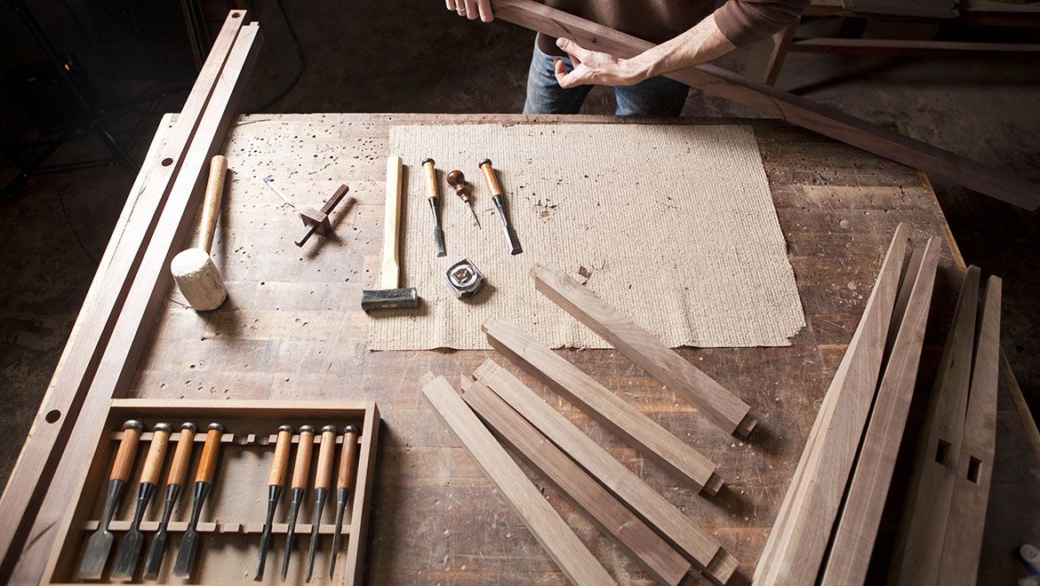 View of a table filled with wooden pieces and hand tools. There is a person standing at the edge of the table holding a piece of wood