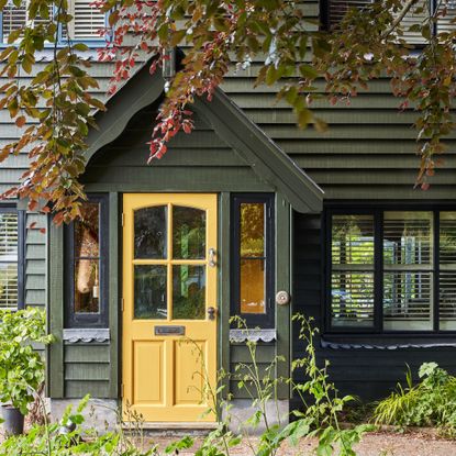 exterior of woodclad house painted dark olive green with yellow front door on porch