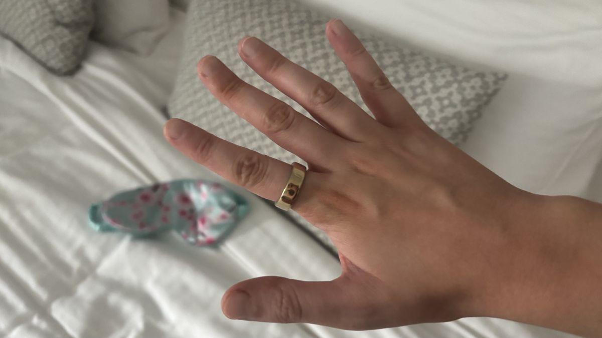 A women's hand models the Oura Ring 5 before a freshly-made hotel bed.
