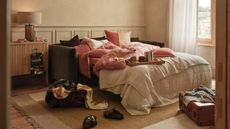 Image of a green velvet sofa bed pulled out into sofa form with off-white and warm pink bedding on it. There is a beige rug underneath the sofa, and two open suitcases in the room.