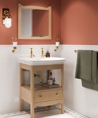 A small bathroom with terracotta and white walls, a rectangular mirror, a wall sconce, and a standing wooden anity with a white basin and gold faucets, with a sage towel next to it