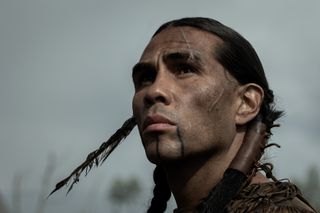 derek hinkey as indigenous character red feater with braids and a feather in his hair as he stares off into the distance in american primeval