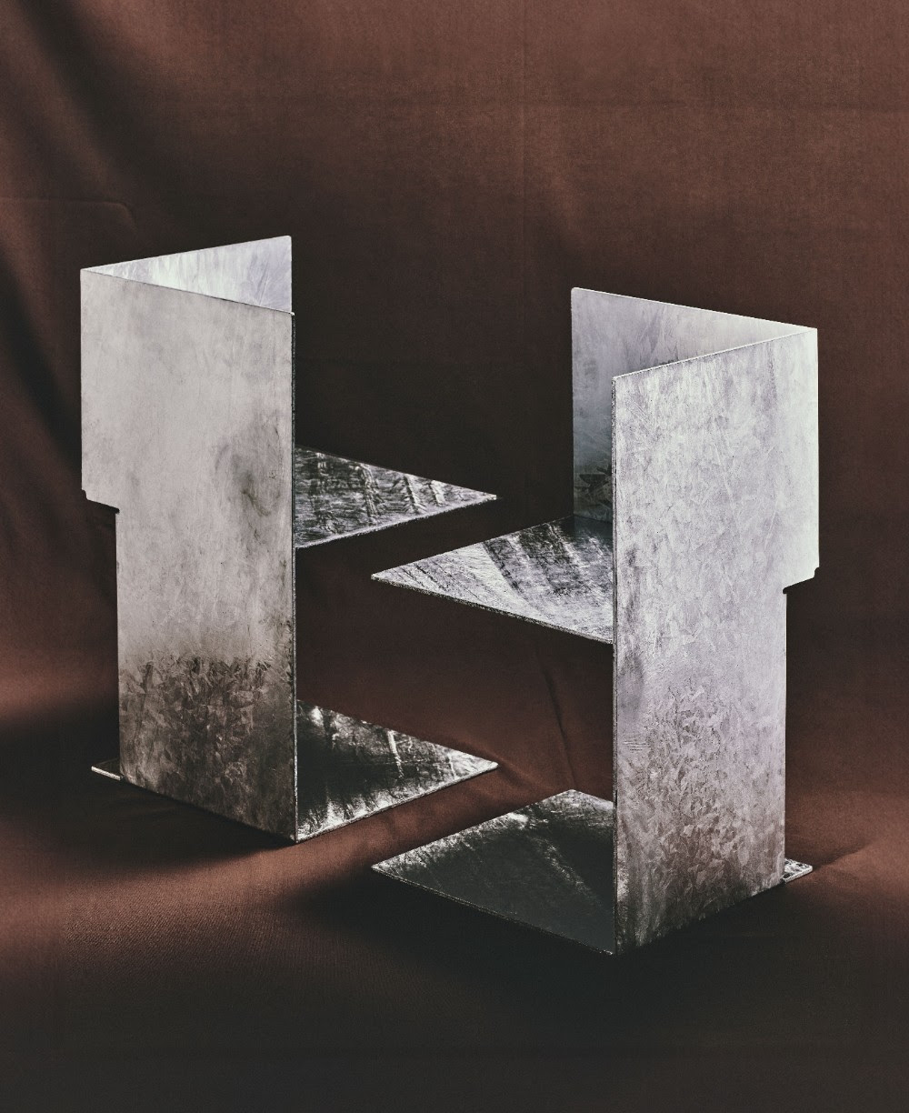 Two sculptural chairs with a super thin, geometric silhouette made of multiple square and rectangular facets sit against a brown background in fabric.