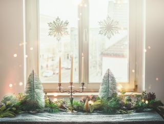 A Christmas decorated table with a candlestick, bottle-brush Christmas trees, and pine cones, in front of a window.