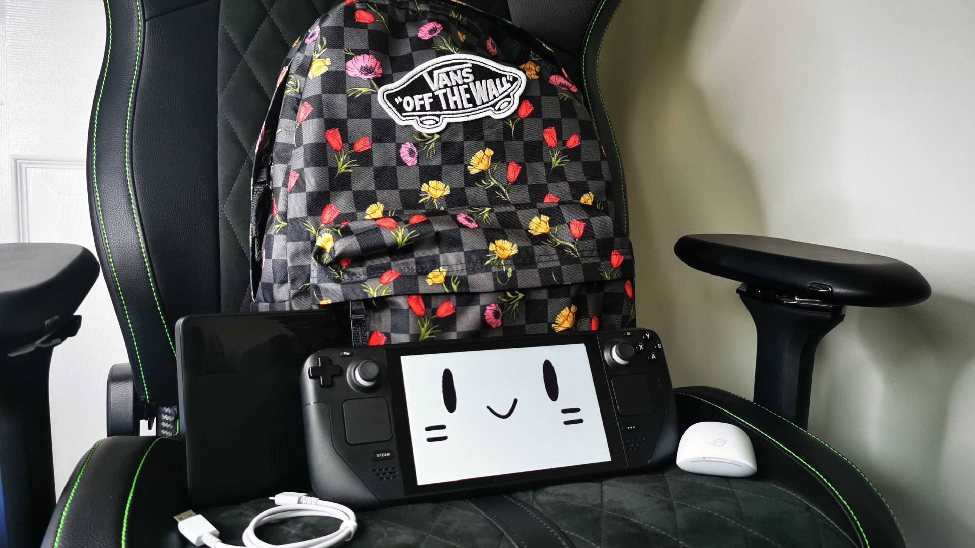 Steam Deck with mascot face on screen sitting on chair with backpack, Asus ROG earbuds, and Baseus power bank