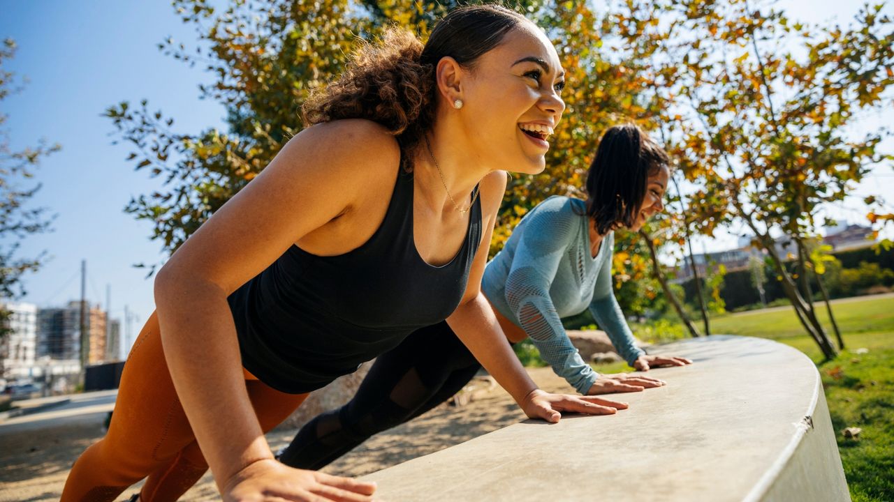 Two women perform push-ups in a park with their hands on a low wall