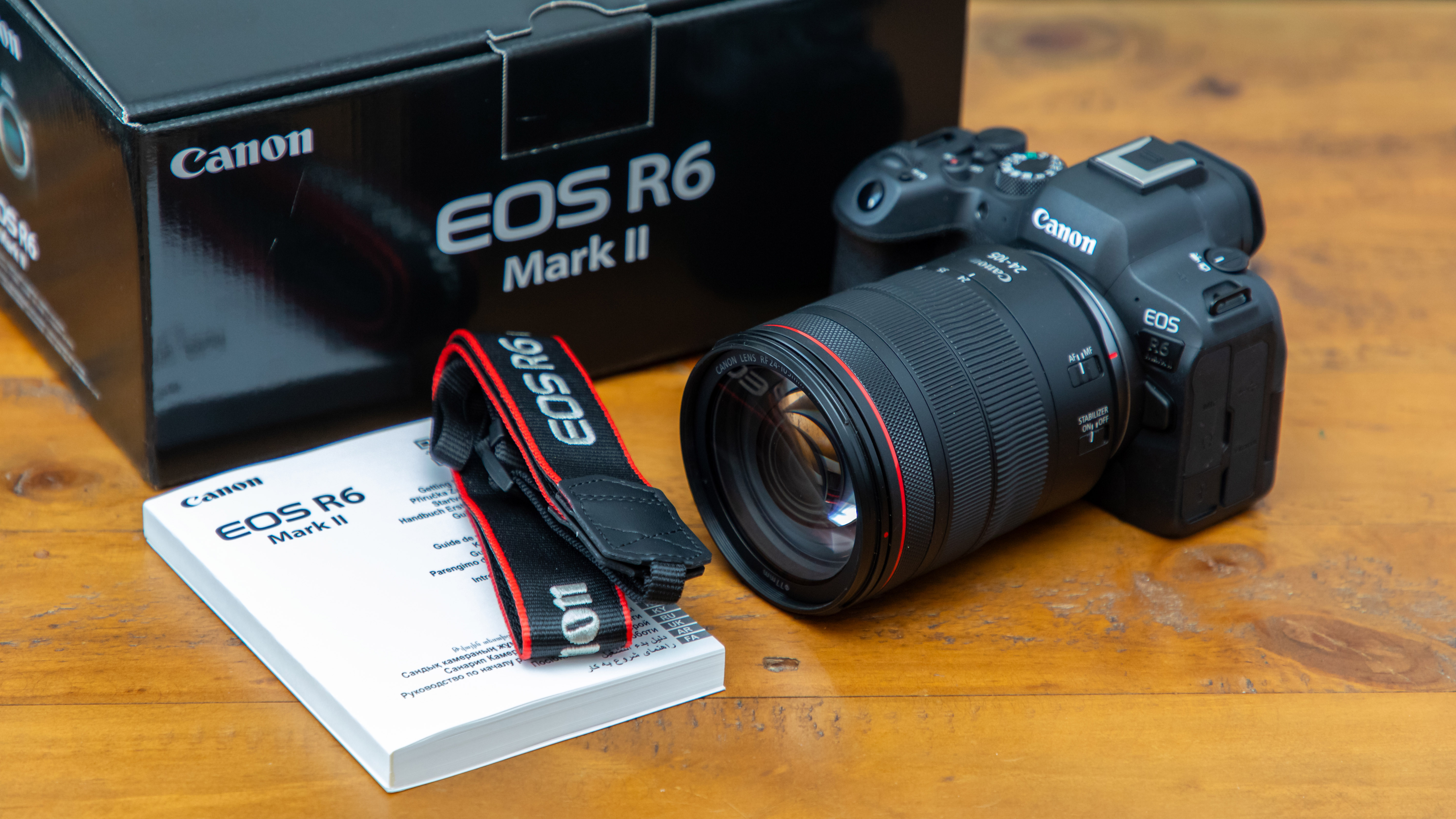 A lifestyle shot of a Canon EOS R6 Mark II on a wooden coffee table next to the camera box, instruction manual and strap neatly placed next to it